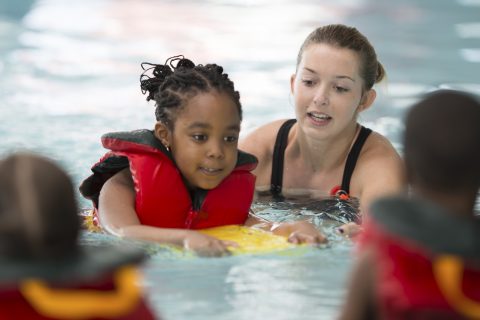 A multi-ethnic group of elementary age children are learning how to swim at the public pool. One little girl is holding onto a kick board and is swimming through the water.