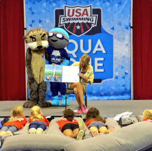 woman and two people in character costumes on a stage reading to children.