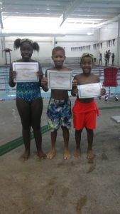 three children holding certificates in front of an indoor pool.