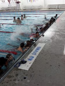 a group of children holding the side of a pool wall and kicking their feet making splashes.