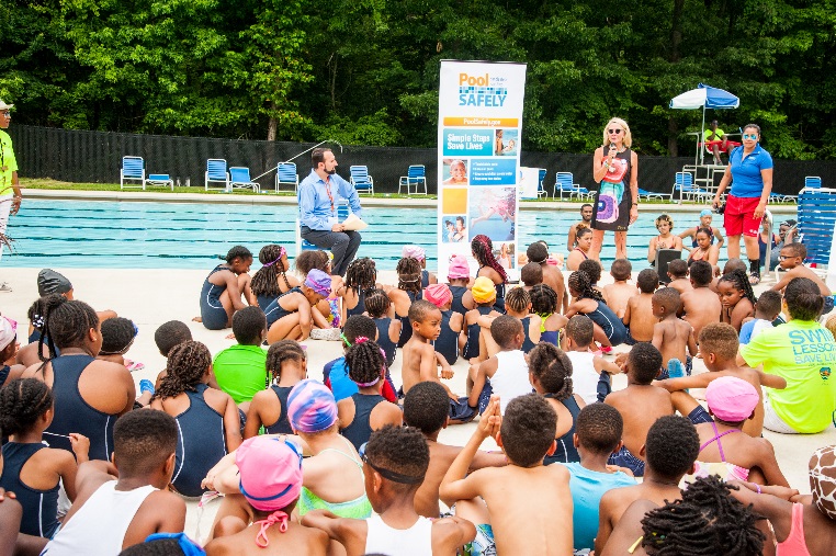 a woman standing in front of a pool speaking to a large group of children.