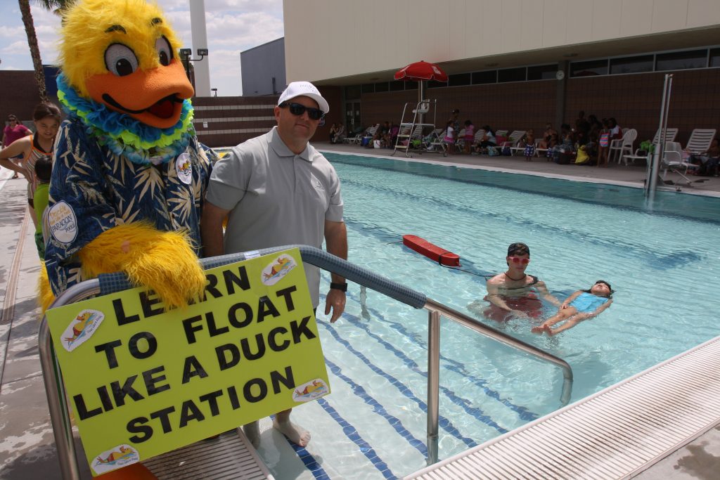 a person in a duck costume standing with a man next to a pool where a lifeguard is helping a girl to float.