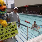 a person in a duck costume standing with a man next to a pool where a lifeguard is helping a girl to float.