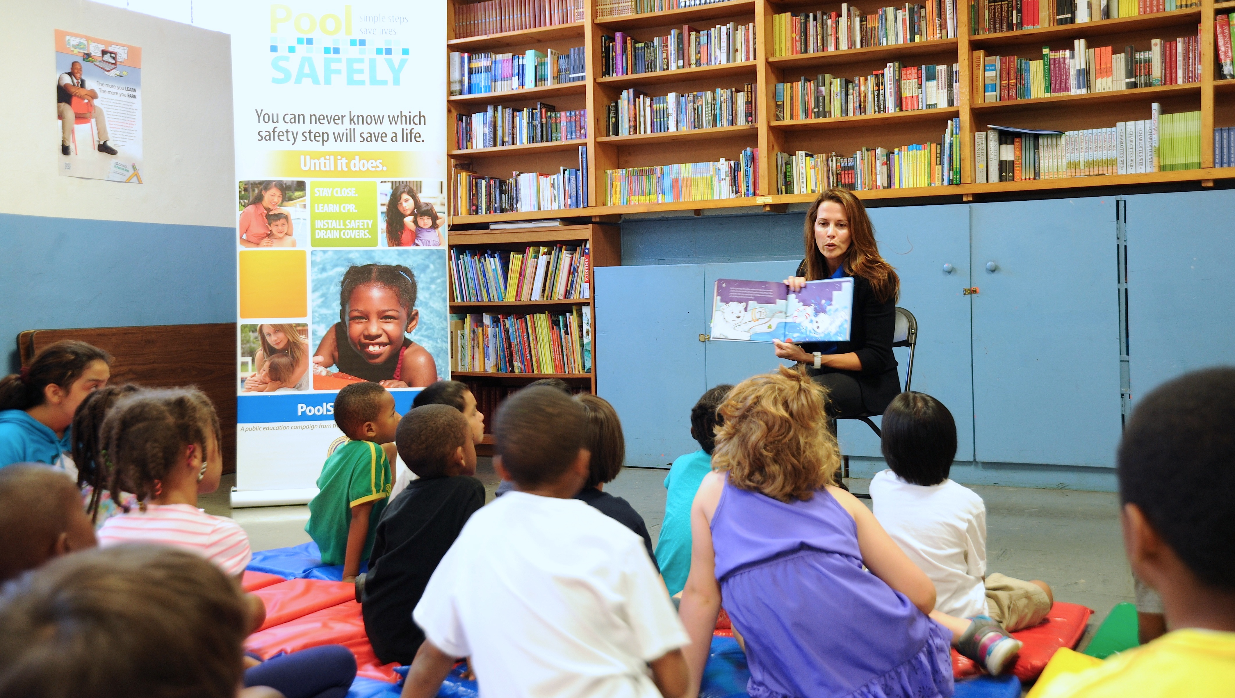 group of children sitting on the floor listening to a woman sitting in a chair reading a book.