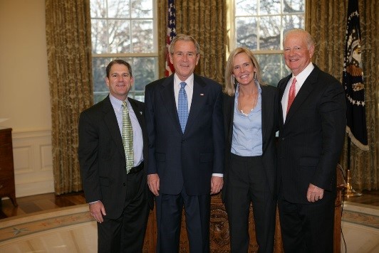 three men and one woman in suits pose for a photo in the oval office at the white house.
