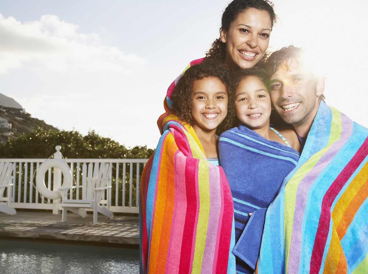 Man and woman with girls in beach towels by pool outdoors.