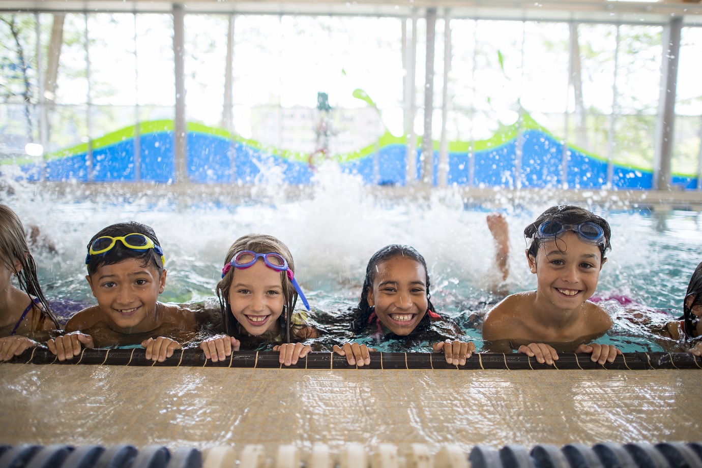 A multi-ethnic group of elementary school children are indoors in a fitness center. They are wearing swimming suits and goggles during swimming class. They are kicking at the side of the pool to practice.