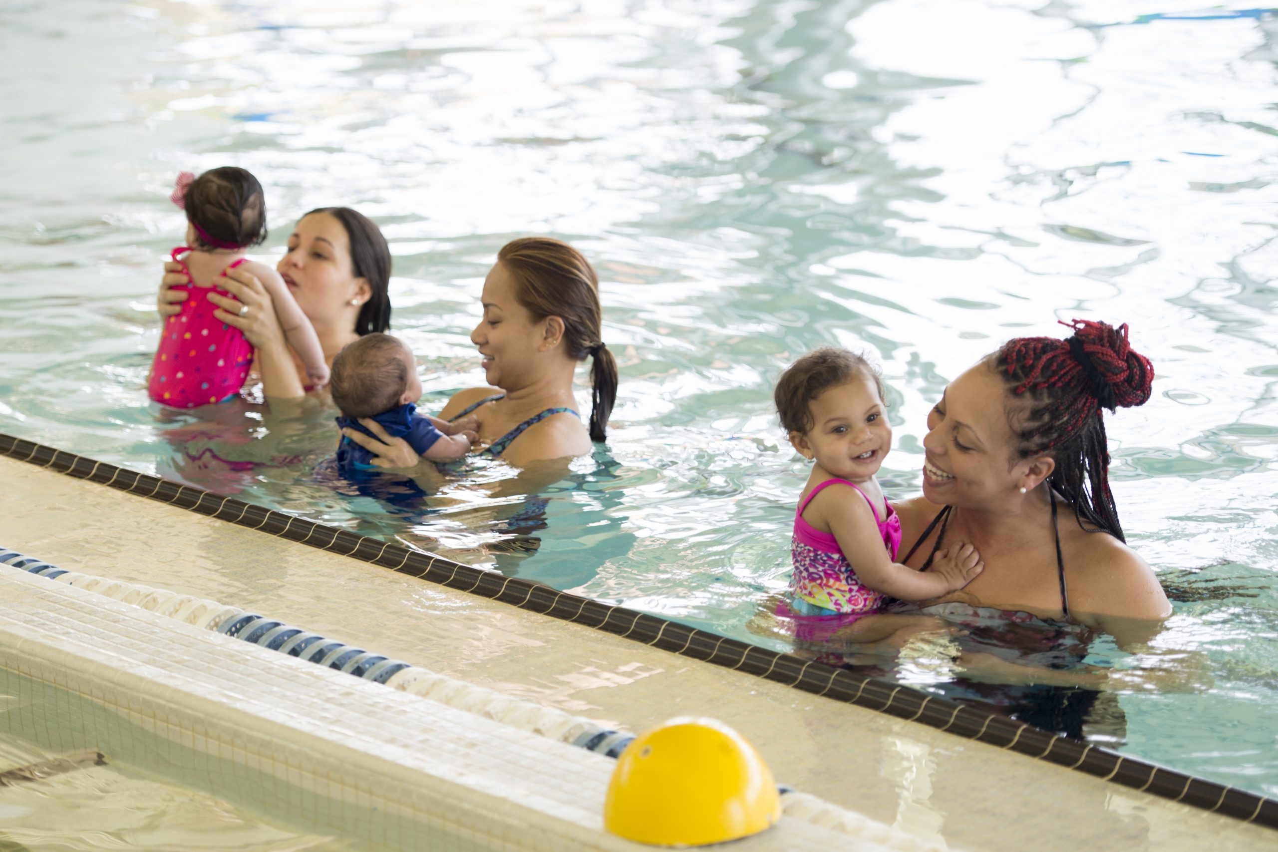 A group of mothers are at the pool and are introducing their young babies to the water at the pool. They are standing in a row and are holding their children.