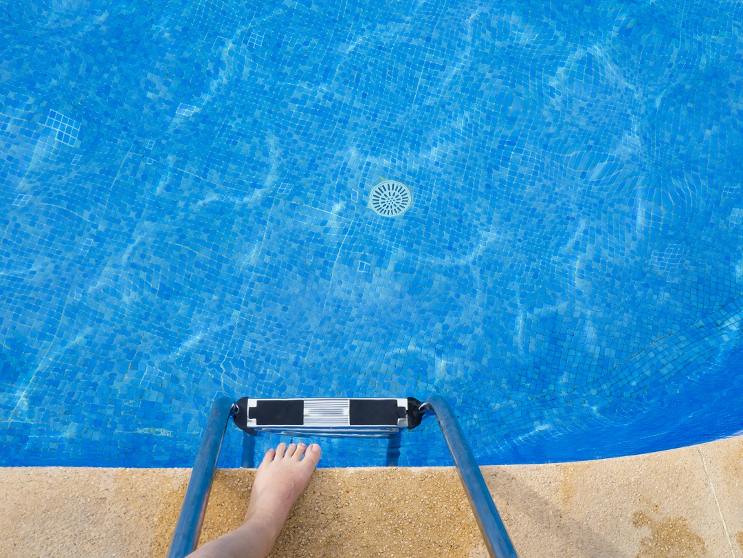 PoV at the edge of a swimming pool, looking down into the water with own feet at the edge of the pool.